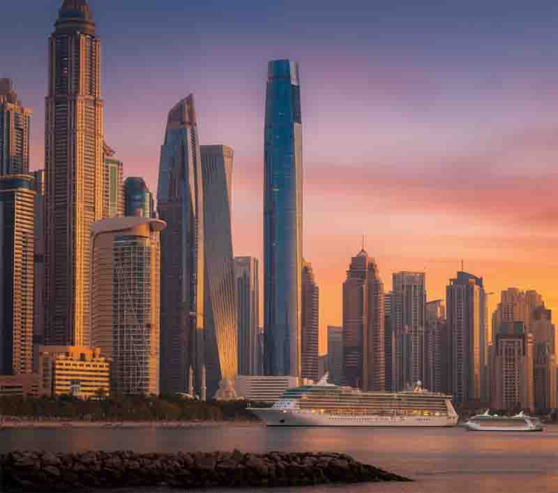 Ciel Dubai Marina Skyline bei Sonnenuntergang in Dubai. Im Vordergrund liegt ein großes, weißes Kreuzfahrtschiff auf dem Wasser, mit einem kleineren Schiff rechts davon. Das Ciel Dubai Marina Hotel dominiert den Hintergrund.