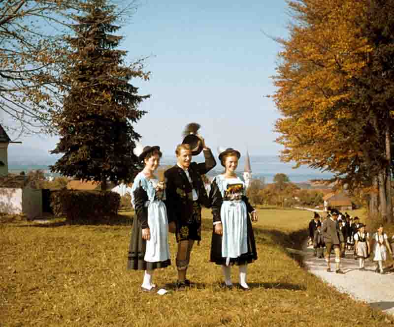 Eine historische Farbfotografie von Ingeborg Bock-Schroeder (Jahrgang 1936) aus den 1950er/60er Jahren in Oberbayern. Das Bild zeigt eine Gruppe junger Menschen in traditioneller bayerischer Gebirgstracht auf einer herbstlichen Wiese. Im Vordergrund stehen zwei Frauen in Dirndln mit hellblauen Schürzen und Hüten mit weißer Feder sowie ein Mann in bestickter Lederhose, der lächelnd seinen Hut mit Gamsbart hebt.