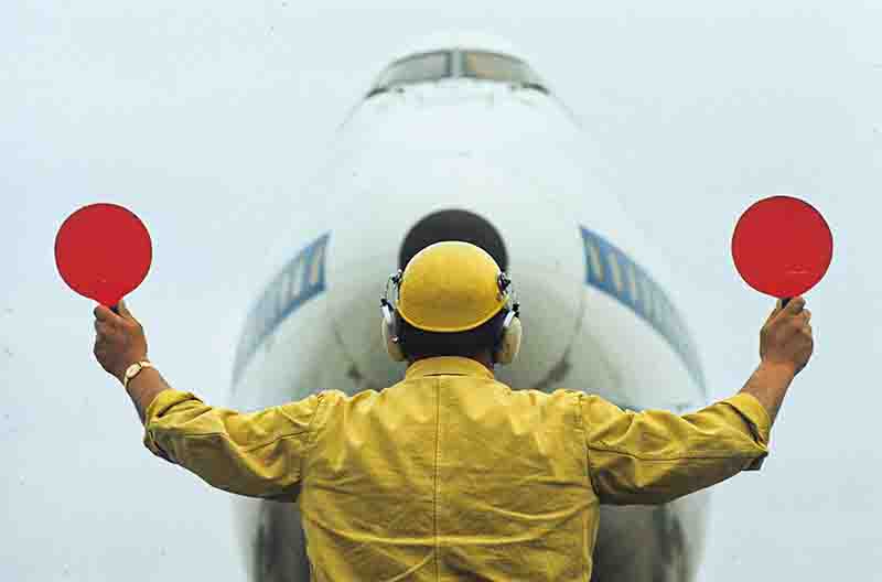 Ein Flugzeug-Einwinker (Marshaller) von hinten gesehen, der in gelber Arbeitskleidung, Helm und Gehörschutz vor der Nase eines großen Verkehrsflugzeugs steht. Er hält beide Arme seitlich nach oben und hält zwei runde, rote Signalkellen hoch, um das Flugzeug einzuweisen.